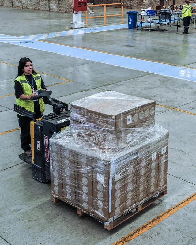 Operator driving an electric pallet jack with palletized cargo inside a warehouse.