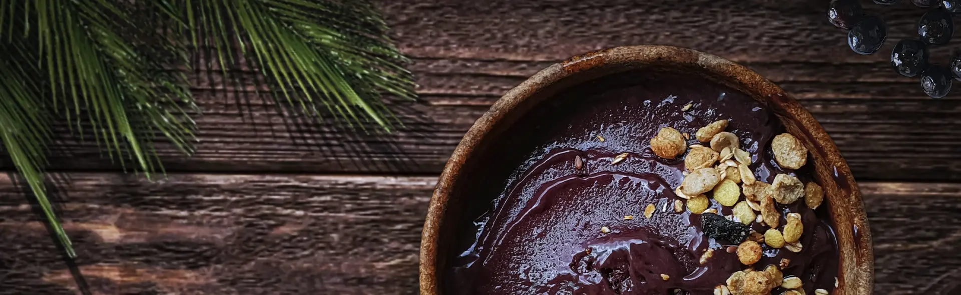 Tigela de açaí com granola sobre mesa de madeira, com folhas de palmeira ao lado.