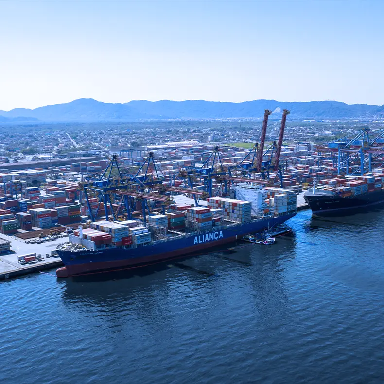 Aliança container ship berthed at a port terminal, with cranes and the city in the background.