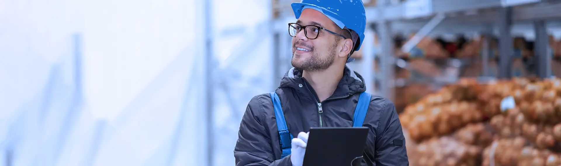 Operador sorridente de capacete azul, com um tablet na mão, dentro de um armazém com sacas de mercadoria ao fundo.
