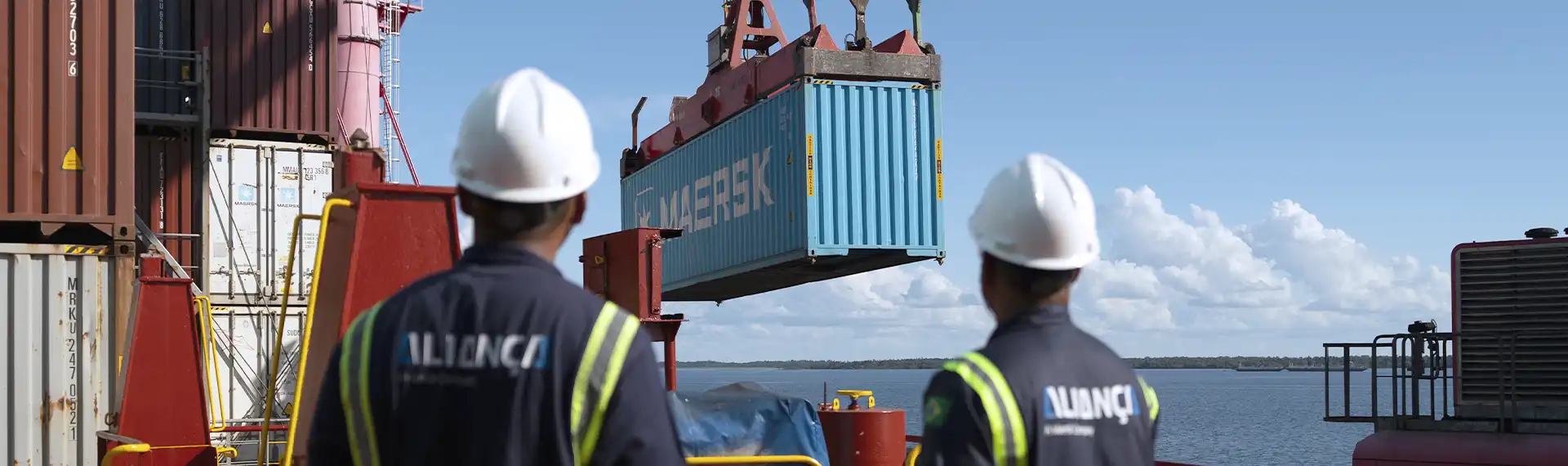 Dois operadores da Aliança acompanham o içamento de um contêiner Maersk a bordo de um navio.
