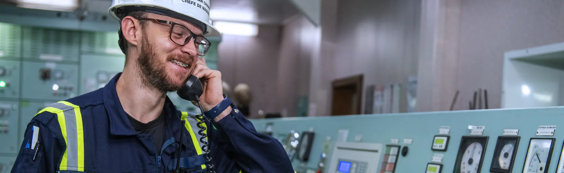 Crew member in a hard hat smiling on the phone in a ship’s control room.