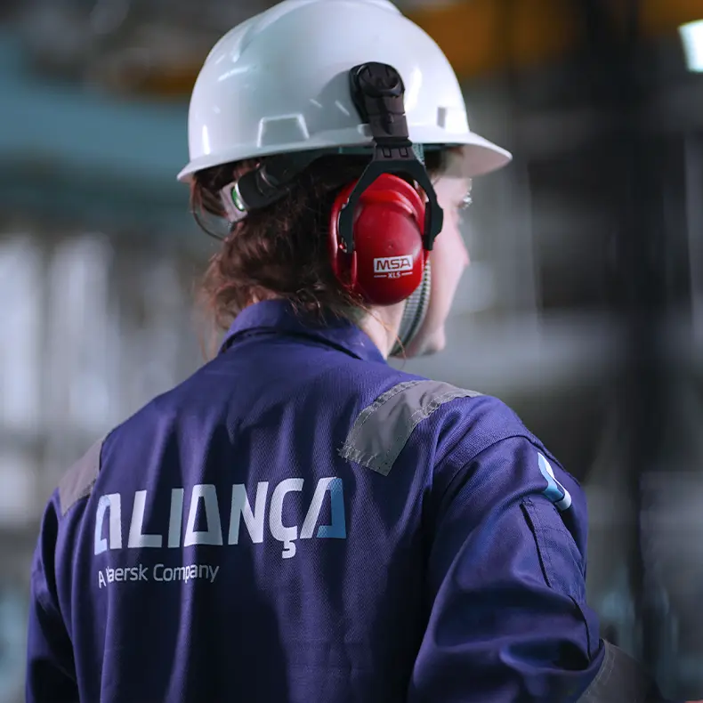 Colaboradora da Aliança de costas, com uniforme azul, capacete branco e protetor auricular vermelho em ambiente industrial.