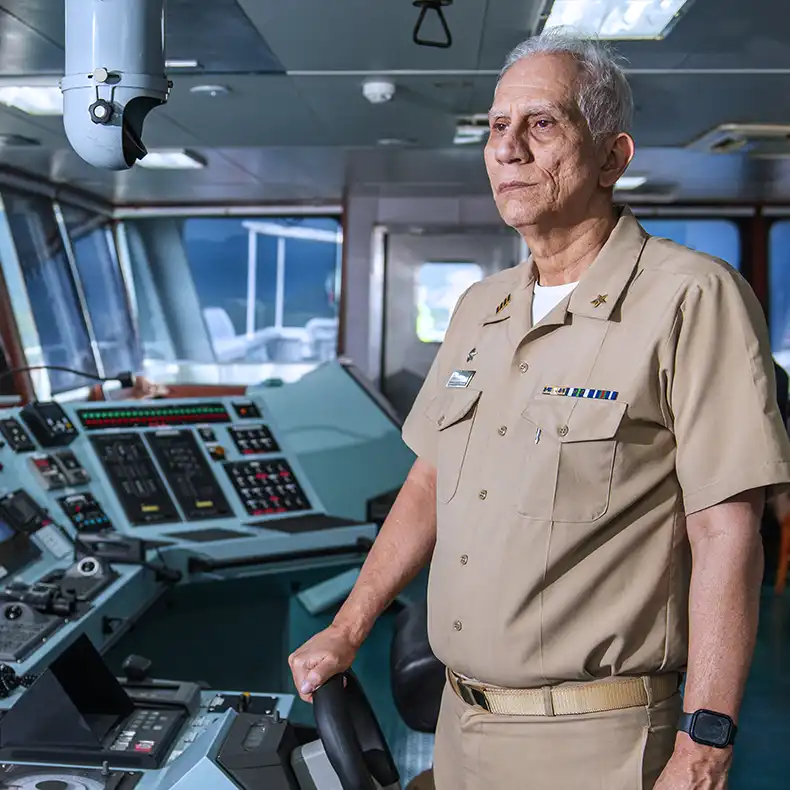Comandante uniformizado no passadiço de um navio, com a mão no timão.
